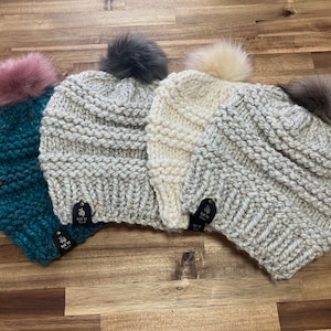 May include: Four chunky knit beanies in teal, light gray, cream, and gray, each topped with a faux fur pom. The beanies have a ribbed knit pattern and small black tags. The hats are displayed on a wooden surface.