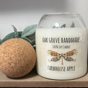 Pode incluir: Uma vela de vidro transparente em um frasco com um rótulo branco que diz "Oak Grove Handmades 100% Soy Candle Farmhouse Apple". O rótulo apresenta um design de libélula colorido.