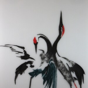 May include: A white ceramic tile with a black and white illustration of two cranes in a dancing pose. The cranes have red accents on their heads. The tile is signed "Ian" in the bottom right corner.