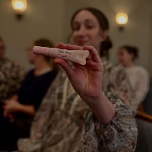 Könnte beinhalten: Eine rosa Lipgloss-Tube mit dem Text "Perfectly Imperfect" und "Soft Champagne" aufgedruckt. Die Lipgloss-Tube wird vor einem verschwommenen Hintergrund gehalten.