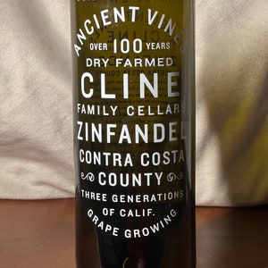 May include: A dark green glass wine bottle with white text that reads "Vintage 2020 Ancient Vines Over 100 Years Dry Farmed Cline Family Cellars Zinfandel Contra Costa County Three Generations of Calif. Grape Growing".