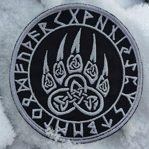 May include: A black and white embroidered patch featuring a bear paw print encircled by a ring of runes. The runes are stitched in white thread on a black background.