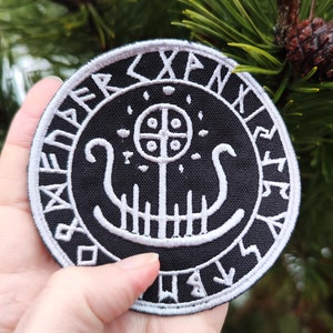 May include: A black embroidered patch featuring a white design of a ship with a tree and a circle with four dots. The ship is surrounded by a circle of runes. The patch is being held in a hand.