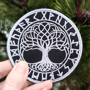 Könnte beinhalten: Ein weiß gestickter Patch mit schwarzem Hintergrund, der ein Lebensbaum-Design zeigt, das von einem Kreis aus Runen umgeben ist. Die Runen sind weiß und lauten "XPNRWYHWA".
