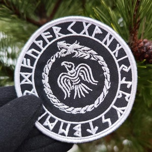May include: A black and white embroidered patch featuring a raven and a snake within a circle. The circle is surrounded by white runes.