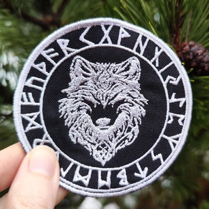 Könnte beinhalten: Ein gestickter Patch in Schwarz und Weiß mit einem Wolfskopf, der von einem Kreis aus weißen Runen umgeben ist. Der Patch ist rund und hat einen schwarzen Hintergrund.