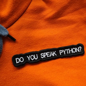 Op de afbeelding: Oranje hoodie met een zwarte patch waarop staat "Do you speak Python?"