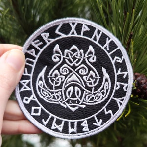 May include: A black and white embroidered patch with a circular design. The center of the patch features a stylized boar's head with intricate knotwork. The outer circle is a band of runes.