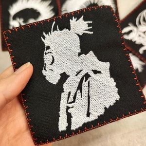 Pode incluir: Um remendo preto bordado com uma silhueta branca de uma pessoa com um coque. O remendo tem costuras vermelhas ao redor das bordas.