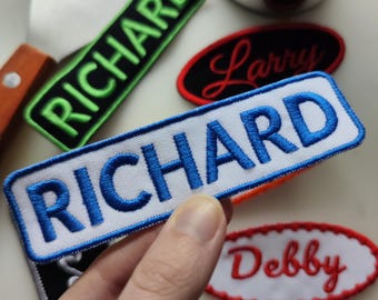 Op de afbeelding: Een hand die een witte rechthoekige patch vasthoudt met de naam "RICHARD" in blauw geborduurd. Andere naam-patches, waaronder "Larry" en "Debby", zijn ook zichtbaar, evenals een groene "RICHARD" patch.