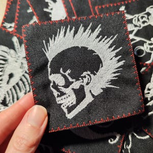 May include: Square black patch with a white skull design and a red stitched border. The skull has a mohawk and is embroidered on the patch. The patch is likely for clothing or accessories.