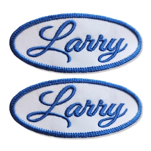 Op de afbeelding: Twee ovale patches met de naam "Larry" geborduurd in blauw script. De patches hebben een witte achtergrond en een blauwe gestikte rand. Het ontwerp is eenvoudig en klassiek, geschikt voor diverse toepassingen.