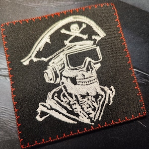 Könnte beinhalten: Weiß gestickter Aufnäher auf schwarzem Stoff mit einem Totenkopf, der einen Piratenhut und eine Schutzbrille trägt. Der Totenkopf hat auf seinem Hut ein Kreuzbein.