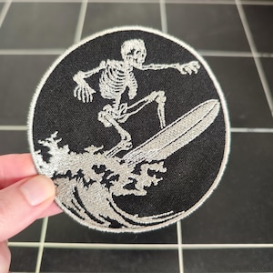 Può includere: Una toppa ricamata in bianco e nero di uno scheletro che fa surf su un'onda. Lo scheletro indossa una tavola da surf bianca e sta cavalcando un'onda con una cresta bianca.