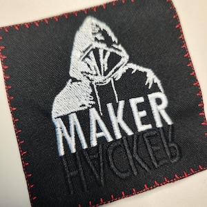 Può includere: Toppa quadrata nera con bordo cucito rosso. La toppa presenta un disegno ricamato bianco di una figura incappucciata e la parola "MAKER" sopra "HACKER" in bianco e nero.