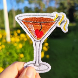 Op de afbeelding: Geborduurde strijkpatch van een cocktailglas met een kers en een citroenschil. Het glas is gevuld met een bruine vloeistof en heeft een witte omtrek.