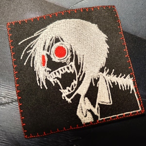 Könnte beinhalten: Weißer gestickter Aufnäher auf schwarzem Stoff mit roten Augen und einem gestickten Rand. Der Aufnäher zeigt einen stilisierten Anime-Charakter mit weit offenem Mund und scharfen Zähnen.