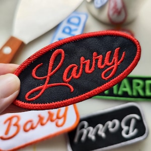 Może przedstawiać: Naszywka haftowana w kształcie czerwonego owalu z imieniem "Larry" w czerwonych literach ozdobnych na czarnym tle.