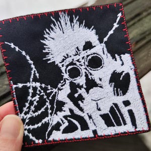 Può includere: Toppa quadrata nera con una grafica bianca di una figura con occhiali e capelli a punta, cucita con filo bianco. La toppa è bordata con cuciture rosse.
