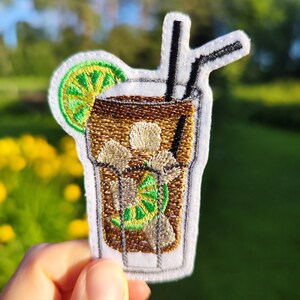 Peut inclure: Patch brodé représentant une boisson glacée brune avec un quartier de citron vert et deux pailles. La boisson est dans un grand verre avec un bord blanc.