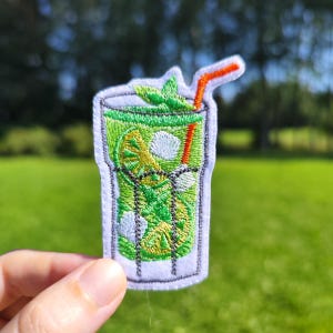 Könnte beinhalten: Gestickter Bügelaufkleber mit einem grünen Mojito-Cocktail mit einer Limettenscheibe, Minzblättern und einem roten Strohhalm in einem Glas.