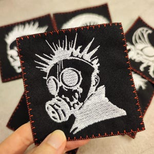 Könnte beinhalten: Weiß gestickter Patch auf schwarzem Stoff mit roten Nähten. Der Patch zeigt eine Person mit Gasmaske und Irokesenschnitt.