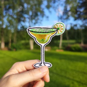 Op de afbeelding: Een geborduurde patch met daarop een margarita-cocktailglas met een limoenwig, in groen en geel. De patch wordt in een hand gehouden.