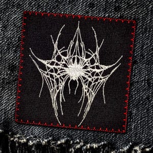 Op de afbeelding: Een zwarte geborduurde patch met een wit spinnenwebontwerp. De patch heeft een rode gestikte rand.