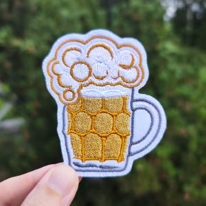 Può includere: Toppa da stiro ricamata bianca e gialla di una boccale di birra con la schiuma in cima.
