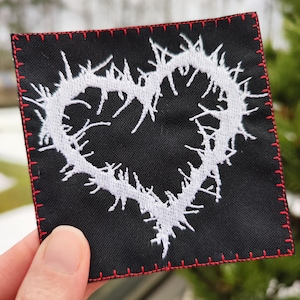 Peut inclure: Patch en tissu noir avec un cœur brodé blanc entouré d'épines. Le patch a des coutures rouges autour des bords.