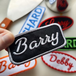 Könnte beinhalten: Ein schwarzer, rechteckiger Aufnäher mit dem Namen "Barry" in weißer Schrift gestickt. Der Aufnäher hat einen grauen Rand und ist von anderen Namensaufnähern in verschiedenen Farben umgeben, darunter "Richard" und "Debby".