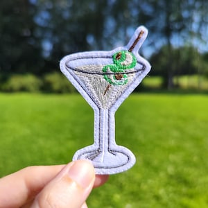 Peut inclure: Patch brodé blanc représentant un verre à martini avec une olive verte et un cure-dent.