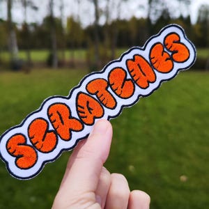 May include: Orange felt patch with the word "SCRATCHES" in a bubble-like font.