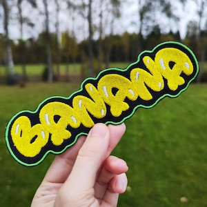 Könnte beinhalten: Ein gelber gestickter Aufnäher mit dem Wort "BANANA" in einer blasenartigen Schrift. Der Aufnäher hat einen schwarzen Hintergrund und einen grünen Rand.