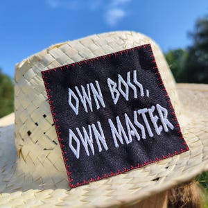 Könnte beinhalten: Strohhut mit einem schwarzen Aufnäher auf der Vorderseite. Der Aufnäher hat einen weißen, gestickten Text mit der Aufschrift "OWN BOSS, OWN MASTER" mit einem rot genähten Rand. Der Hut ist aus geflochtenem Stroh.