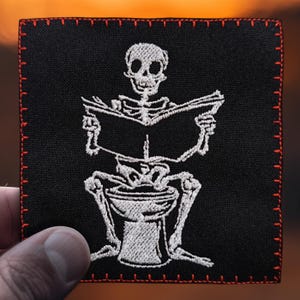 Op de afbeelding: Een wit geborduurde patch op zwarte stof die een skelet toont dat op een toilet zit en een boek leest. De patch heeft rode stiksels rond de randen.