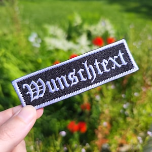 Può includere: Una toppa rettangolare nera con testo ricamato bianco "Wunschtext" in carattere gotico. La toppa ha un bordo bianco. Lo sfondo è una vista sfocata di fogliame verde e fiori rossi.