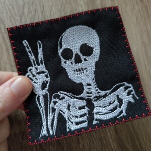 Könnte beinhalten: Weiß gesticktes Skelett mit einem Peace-Zeichen auf einem schwarzen Stoff-Patch mit roten Nähten an den Rändern.
