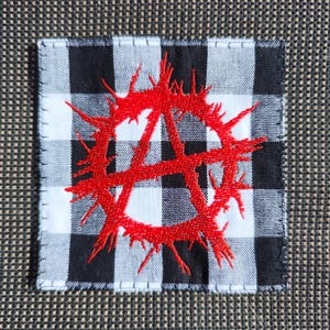Può includere: Un cerotto di stoffa a quadri bianchi e neri con un simbolo anarchico ricamato in rosso.