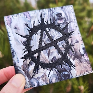 Könnte beinhalten: Ein weißer Stoff-Patch mit einem gestickten schwarzen Anarchiesymbol, umgeben von einer Collage aus grauen und weißen Hundefiguren.