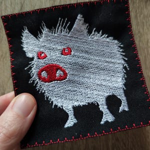 Peut inclure: Patch brodé blanc représentant un cochon de dessin animé avec des yeux et un groin rouges sur un fond de tissu noir. Le patch a une bordure cousue rouge.