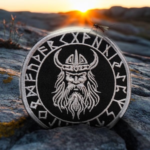 May include: A black and white embroidered patch featuring a Viking warrior wearing a horned helmet. The patch is encircled by a ring of runes. The patch is resting on a rocky surface.