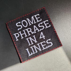 Può includere: Toppa quadrata nera ricamata con la scritta "SOME PHRASE IN 4 LINES" in bianco. La toppa ha un bordo cucito rosso. La toppa è su una superficie scura.