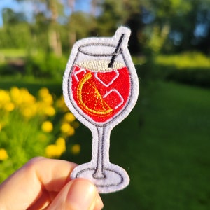 Op de afbeelding: Een witte geborduurde patch met een glas rode wijn met een sinaasappelschijfje en een rietje. Het glas staat op een witte achtergrond.