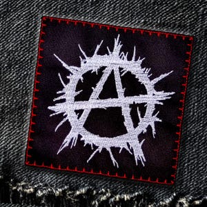 Op de afbeelding: Een zwarte stoffen patch met rode stiksels rond de randen. De patch heeft een wit anarchie symbool met een cirkel van spikes eromheen.
