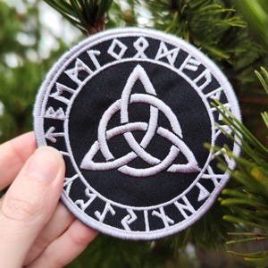 Könnte beinhalten: Ein gestickter Patch in Schwarz und Weiß mit einem weißen keltischen Knotendesign in der Mitte. Der Knoten ist von einem Kreis aus weißen Runen umgeben.