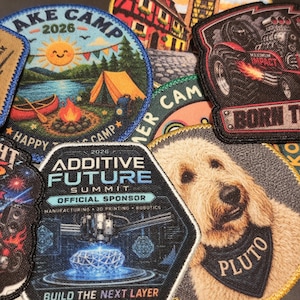 Könnte beinhalten: Eine Sammlung bestickter Aufnäher mit verschiedenen Motiven, darunter eine Lake Camp 2026-Szene mit Zelt und Lagerfeuer, ein Maximum Impact Monstertruck, ein Additive Future Summit 2026 Sponsoren-Aufnäher und das Porträt eines Hundes mit Pluto-Halstuch.