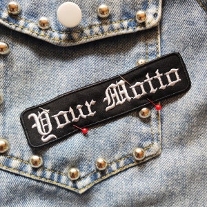 Op de afbeelding: Zwarte stoffen patch met witte geborduurde tekst die "Your Motto" zegt op een blauwe denim jas met zilveren studs.
