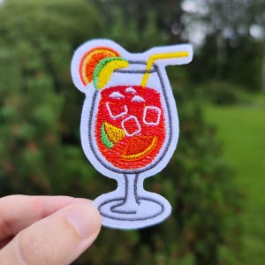 May include: Embroidered iron-on patch of a red cocktail with ice cubes, a yellow orange slice, and a green lime wedge. The cocktail is in a clear glass with a white stem and base. A yellow straw is sticking out of the top of the glass.