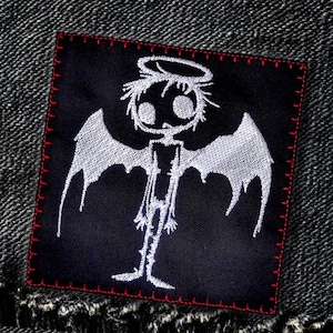 以下が含まれることがあります： 黒い布のパッチに、白い刺繍で翼と後光を持つ骸骨の姿が描かれています。その姿は、大きな目をした頭蓋骨のような頭をしています。